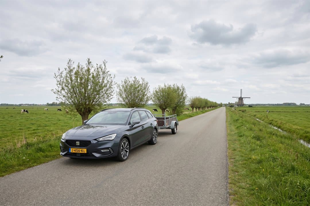 Auto met open aanhanger op een polderweg, met molen en knotwilgen op de achtergrond